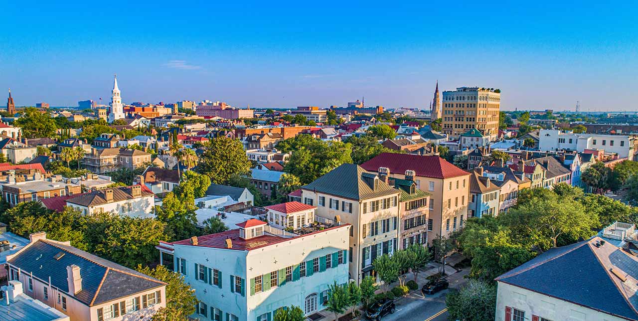 An elevated panoramic view of a historic city featuring colorful multi-story buildings, lush green trees, and prominent church steeples under a clear blue sky