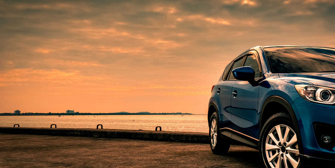 A blue SUV parked on a pier overlooking a calm body of water under a warm, golden sunset sky