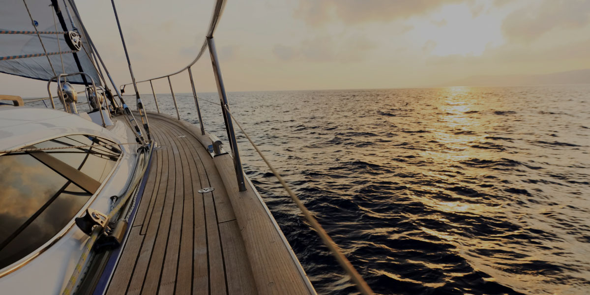 View from the wooden deck of a sailboat moving across dark water toward a golden sunset on the horizon