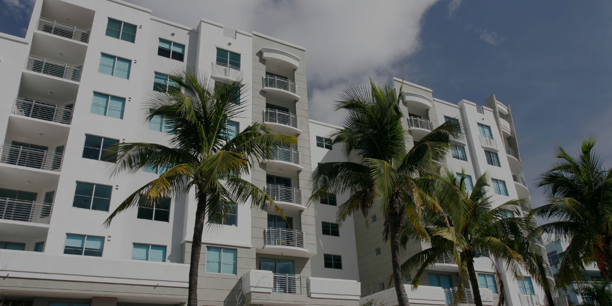 A low-angle view of a modern white apartment building with multiple balconies and large windows, framed by several lush palm trees under a partly cloudy sky