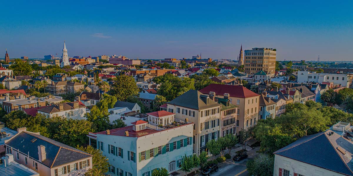 An elevated panoramic view of a historic city featuring colorful multi-story buildings, lush green trees, and prominent church steeples under a clear blue sky