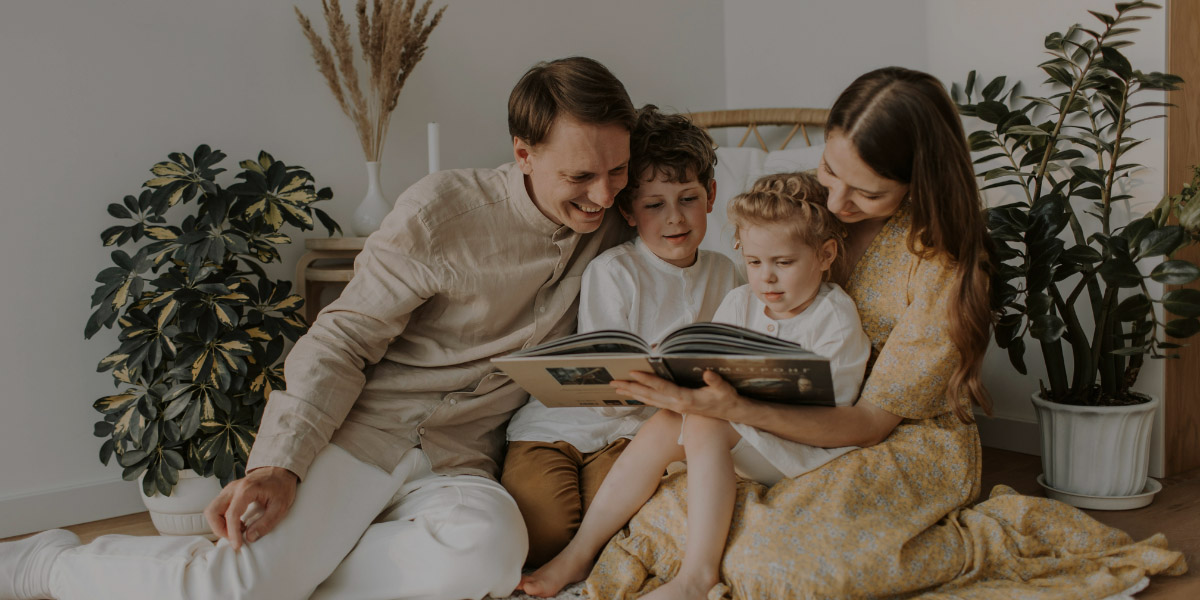 A smiling family of four sits together on the floor, reading a book in a cozy room filled with indoor plants