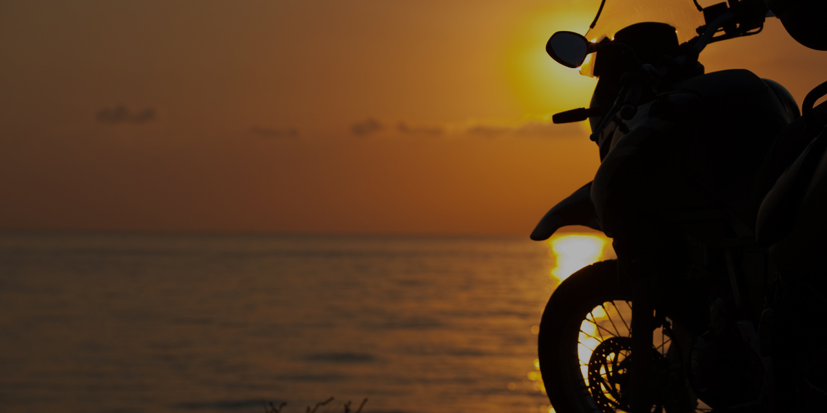 Silhouette of a motorcycle parked by the sea during a golden sunset, with the sun reflecting on the calm water