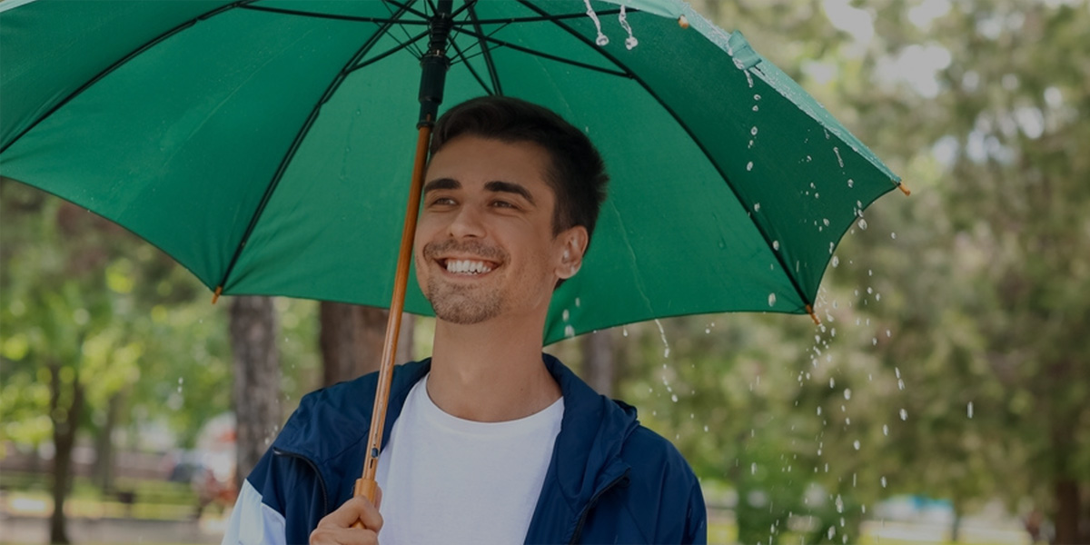 A smiling young man stands outdoors under a large green umbrella during a rain shower