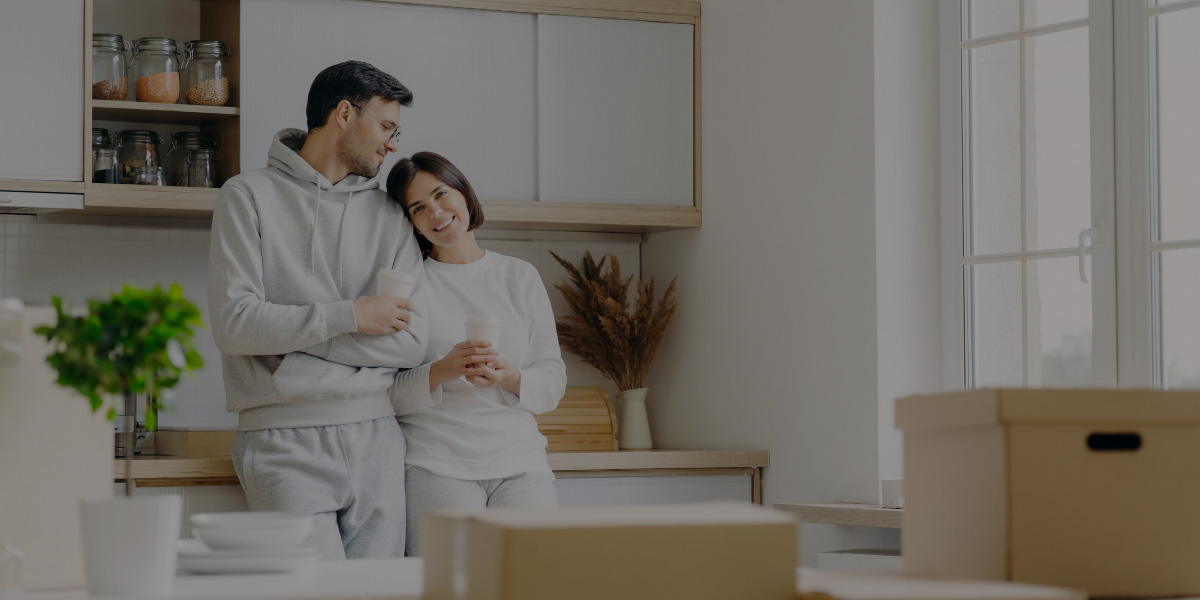 A smiling couple in comfortable grey loungewear stands together in their new kitchen, holding coffee cups amidst unpacked moving boxes